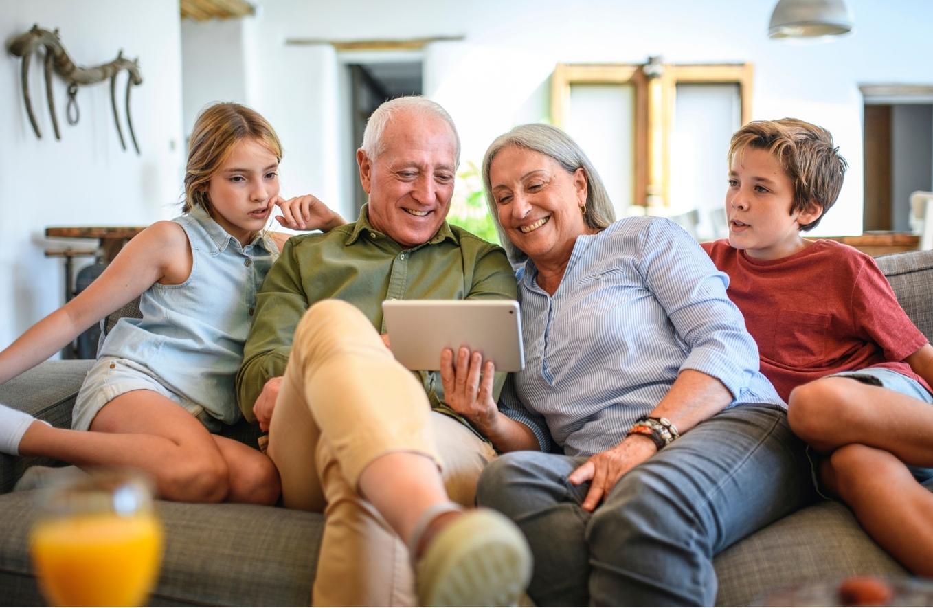 Family of four people sitting on couch. Grandparents holding tablet computer with grandchildren either side.