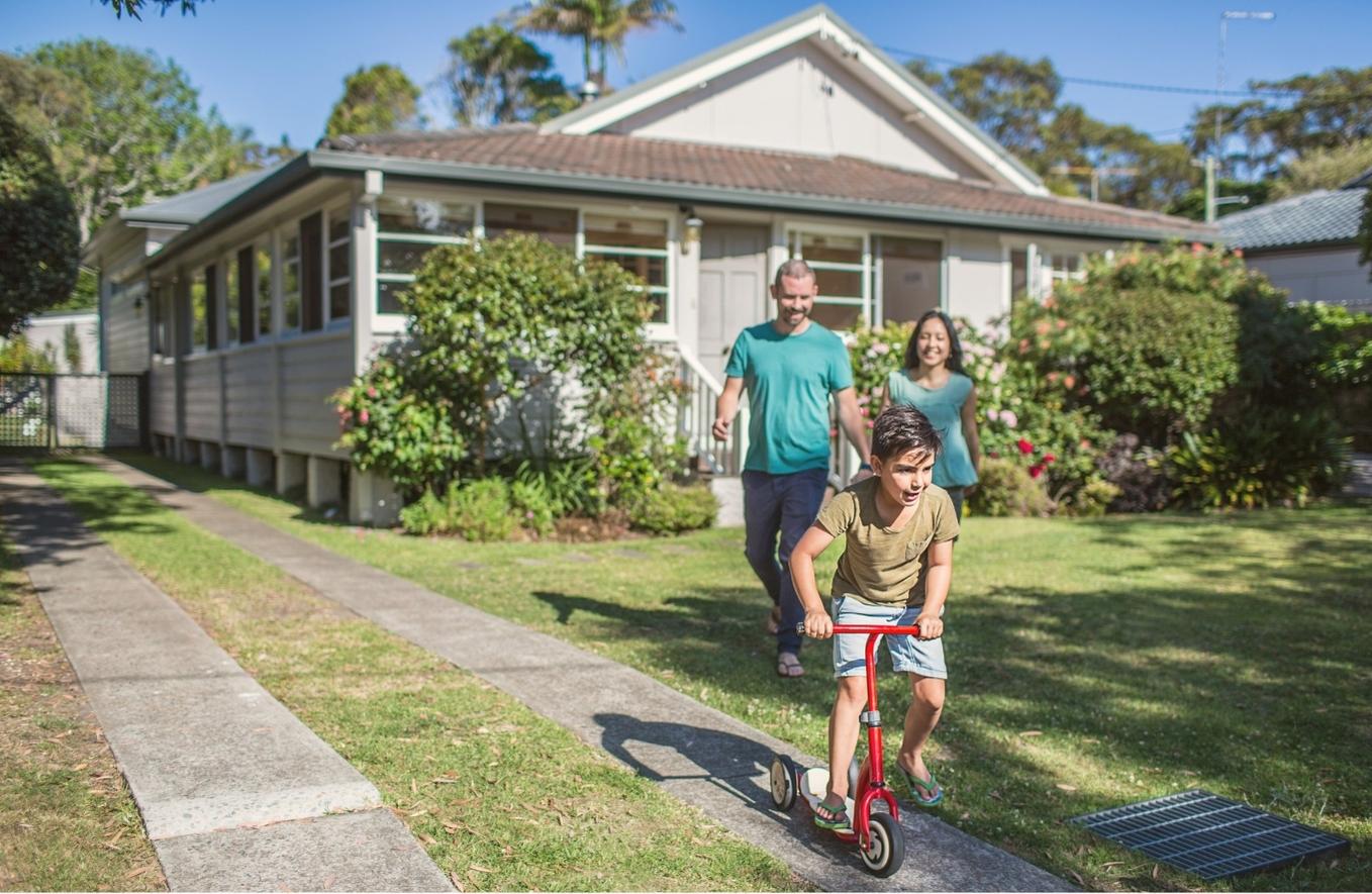 Boy riding on red scooter in front yard with parents watching